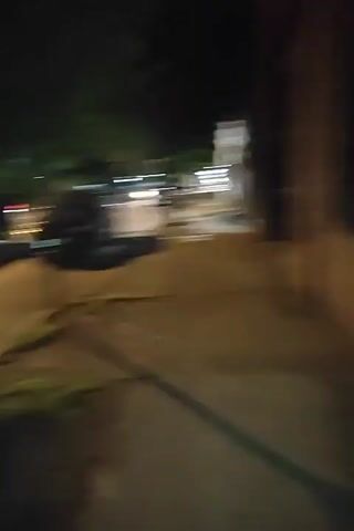 Here is a detailed description of the video:

The video begins with a blurred view from inside a car at night. The dashboard lights are visible, and the lights of other cars streak by. The camera abruptly shifts to a man standing outside at night. He is wearing a green jacket, a black baseball cap, and has long hair pulled back in a ponytail. He speaks to the camera, then the camera pans to the right, showing a street at night. There are buildings lining the street, lit by streetlights. A car is parked on the side of the road in front of a building that has a sign on it that reads "Trowley Market." The overall environment is urban and dark.