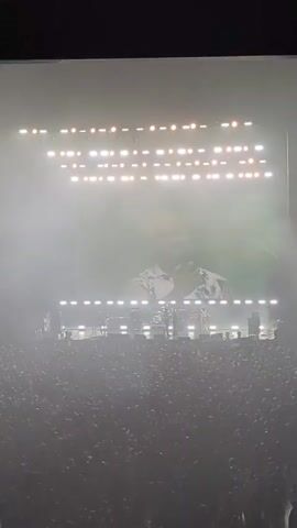 A video clip shows a massive concert crowd filling a stadium. The stage is brightly lit with numerous spotlights and large screens displaying the performers. On the main screen, a man in a patterned shirt is shown, gesturing and interacting with the audience. The atmosphere is energetic and filled with stage fog.