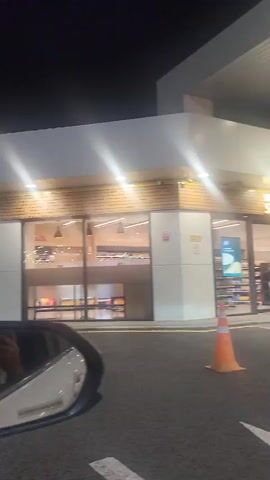The video pans from left to right showing the exterior of a brightly lit convenience store at night, possibly a gas station store, as indicated by the presence of orange traffic cones. The store has large glass windows displaying shelves stocked with various products. Inside, a person can be seen reflected in the side mirror of a car, taking a photo. The reflection shows a person holding a phone up to take a picture. The car appears to be parked in front of the store. The lighting is artificial and reflects off the wet pavement. The sky is dark, indicating nighttime.