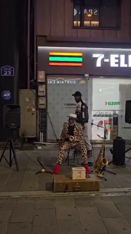 A street performer, an elderly Black man with a white hat, a leopard-print suit, and red shoes, plays a saxophone while seated on a suitcase on a sidewalk at night. Another Black man in a black and white jacket and grey pants stands to his left, seemingly observing. A second saxophone leans against a stool to the performer's right. Two speakers are visible, one on a tripod to the left and another to the right, behind the performer. Behind them is a storefront with a "7-ELEVEN" sign and a rainbow-colored rectangle sign. A street sign with the number "23" is on the brick wall to the left. The ground is paved with cobblestones. The lighting is artificial and casts shadows on the ground.