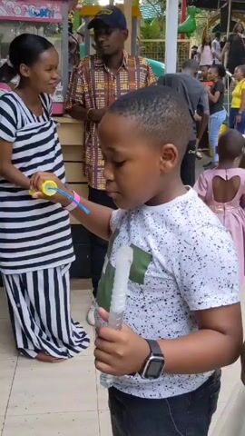 The video captures a moment at an outdoor amusement park, focusing on a young boy engrossed in blowing bubbles. He is positioned at the forefront, wearing a white T-shirt speckled with a pattern, dark pants, and a digital watch. In his hands, he holds a bubble wand and a container of bubble solution, directing his attention to creating bubbles.

Behind him, a woman stands out in a striped black and white outfit, engaged in a conversation with a man in a patterned shirt and a baseball cap. The setting is a bustling environment, with other individuals milling around, including children in party dresses and casual attire. A popcorn stand is visible in the background, adding to the lively ambiance of the park.