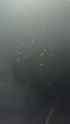 The video appears to show fireworks being launched at night. Initially, there is a dark gray background with a few small orange specks, possibly embers or the very beginning of a firework. Then, the scene transitions to a completely black background as fireworks are launched and explode near the bottom of the frame. There are flashes of orange sparks and white smoke that dissipate and drift upwards. A long, thin, angled object is visible at the bottom right, possibly the launch tube or a stick attached to the firework. The video mainly focuses on the pyrotechnic display with the background obscured by darkness and smoke.