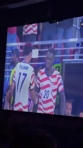 A television screen displays a soccer game in progress. The screen shows a close-up of two players wearing US national team jerseys. One player, number 17, is identified as "TILLMAN." The other player, number 20, has the word "FIFA" superimposed over his face. The game appears to be taking place at a stadium with spectators in the background. The field is green with white lines, and there are various markers and players scattered across it. The scoreboard indicates the score is 0-0. A banner at the top of the screen reads "THE FINAL HALF TIME SHOW."