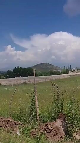 The video shows an outdoor rural scene. In the foreground, there is dirt, dry grass, and a wooden post. In the midground, there is a grassy field with scattered trees and a dirt pile. Further in the background, there are hills, a building with a slanted roof, and a blue car parked on a hill. The sky is blue with scattered white clouds. The camera pans from left to right, capturing different parts of the landscape.