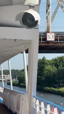 The video shows a view from the deck of a white boat, moving along a canal. A security camera is mounted on the side of the boat. In the background, a train bridge with a clearance sign indicating 14.0 meters is visible. The canal banks are lined with green trees. A person is standing on a paddleboard in the canal. The sky is blue and clear.