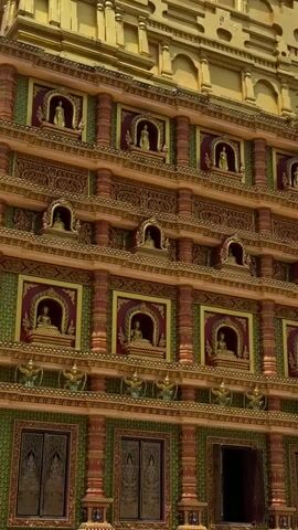 A slow upward pan reveals the ornate golden and green facade of a Buddhist temple. The architecture is highly detailed, with rows of small niches, each containing a golden statue of Buddha. Red and gold decorative elements frame these niches, and intricate patterns adorn the walls. Balconies and decorative pillars add to the visual complexity. The upper portion of the building, also gold, features tiered spires and intricate carvings, suggesting immense scale and craftsmanship. The sky is visible in the background, with some clouds suggesting daylight. No people or active events are present in the frame.