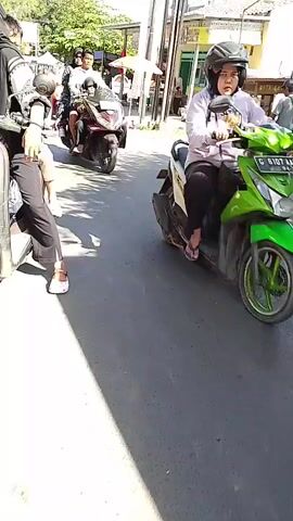 The video displays a street scene with multiple motorcycles and people. The perspective appears to be from a motorcycle moving through the street. In the foreground, shadows of trees are cast on the asphalt road. Several people are visible on motorcycles, some riding in groups, while others are standing or walking on the side of the road. On the right side of the street, there are shops or stalls with people gathered around them. The environment appears to be a sunny day, possibly in a village or a town. The motorcycles are varied in type and color, and some are carrying passengers. The people are dressed in casual clothing, with some women wearing headscarves. The overall impression is of a bustling street with typical daily activity.