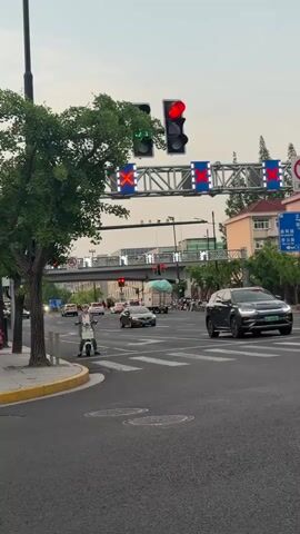 The video begins from the first-person perspective of a motorcyclist facing a red light at an intersection. In the background, there are traffic lights overhead with illuminated red “X” symbols. After the motorcycle stops, the camera focuses on the intersection. There are cars and a truck traveling through the intersection. A person on a small electric scooter waits at the crosswalk for the light to change. They are wearing a helmet and a business casual outfit. A pedestrian overpass spans the street, displaying animated arrows. The scene conveys a normal urban environment with regular traffic flow.
