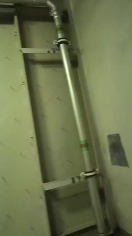 The video shows a section of an industrial interior. The primary focus is a series of vertical metal panels or partitions, possibly part of a larger structure like ductwork. The panels are light-colored, likely painted a pale yellow or off-white. There are faint markings or scratches scattered across the surface of the panels, along with some darker smudges and a spot of pink paint or ink. A white pipe runs horizontally across the top of the frame, partially obscured by the panels. The lighting is dim, contributing to the slightly grimy appearance of the scene. The camera angle and focus suggest a close-up perspective.