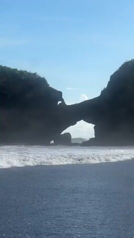 The video shows a beach with black sand. Waves are rolling in.  A rock formation with a large arch in it is in the background, through which the ocean is visible. Greenery can be seen on top of the rock formation. The sky is blue with a few clouds.