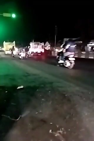 A group of motorcyclists are riding on a road at night. A traffic light is visible, and it is green. There are other vehicles on the road, including trucks. The road surface appears wet. The video is shaky, suggesting it was filmed from a moving vehicle.