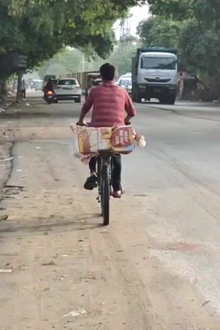 The video shows a man riding a bicycle on a dirt and asphalt road. He is wearing a striped red and white shirt and black pants. The bicycle has a large basket on the back, filled with what appears to be bread or similar packaged goods. He is riding away from the camera, in the middle of a road with other vehicles and trees in the background. The road appears to be a two-lane road with traffic in both directions. There are trees lining the left side of the road and a concrete barrier on the right. Other vehicles visible include cars, a truck, and a motorcycle. The lighting suggests it is daytime.