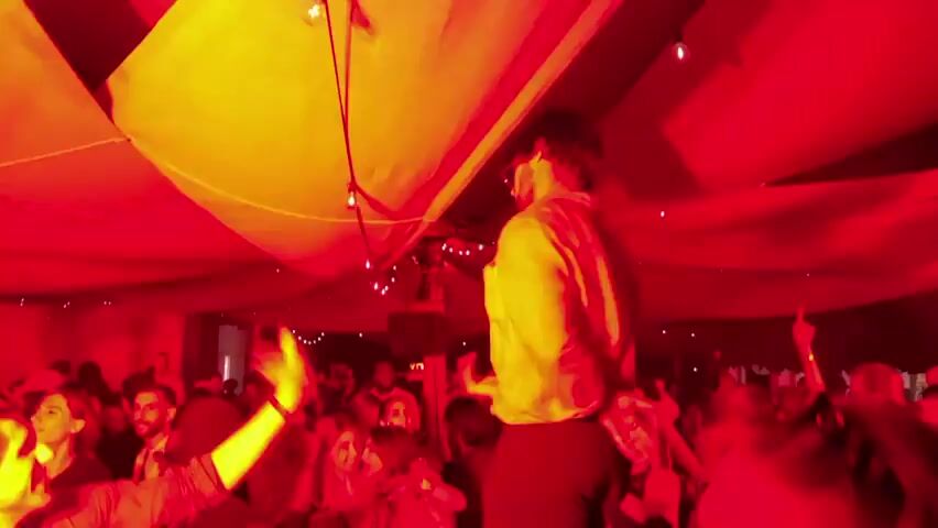A group of people are gathered in a dimly lit room with red lighting. A person wearing a white top and dark pants is standing on what appears to be a platform or elevated surface, looking down at the crowd. The person is gesturing with their hands. The crowd below is diverse and appears to be dancing or moving to music. There are string lights hanging from the ceiling. The environment appears to be an indoor venue, possibly a club or party setting.