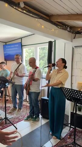 A music band is performing live in a cozy room. Five individuals make up the band: three men and two women. The first man, on the left, plays an electric guitar and wears a white t-shirt and blue jeans. The second man plays a bass guitar, and he is dressed in a white t-shirt and blue jeans with green sneakers. In the center is a woman singing into a microphone, wearing a beige blouse and teal wide-leg pants. Next to her, to the right, a woman sits on a wooden stool playing an acoustic guitar. She wears a white top, a plaid shirt, and blue jeans. The last member, a man to the right, plays an electric drum set.

Behind the musicians, there is a screen showing lyrics in Russian. String lights hang from the ceiling, adding a warm ambiance to the room. The walls are white brick, and there is a window providing natural light. A rug with an oriental pattern covers the floor. There are music stands in front of the singers and guitarist. The room appears to be a cafe or small venue.
