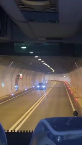 The video is shot from inside a vehicle, likely a bus, looking forward through the windshield. The vehicle is traveling through a tunnel. The tunnel walls are curved and illuminated with yellow and white lights. There are red and blue lights along the side of the road. In the distance, headlights of another vehicle are visible. The interior of the bus is visible in the foreground, including seats and the ceiling. The lighting suggests it is nighttime or the tunnel is artificially lit.