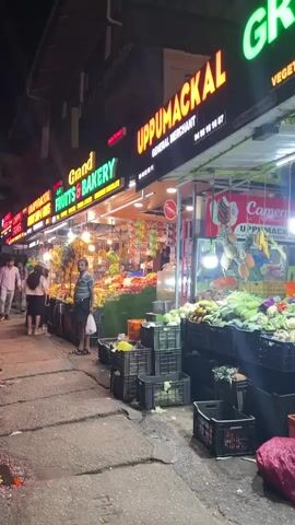 The video shows a night market scene with multiple storefronts displaying fresh produce. In the foreground, numerous black and orange crates are stacked on the sidewalk, overflowing with various fruits and vegetables including leafy greens, gourds, and bananas. A red mesh bag is visible on the ground. Illuminated signs overhead read "UPPUMACKAL," "Good FRUITS & BAKERY," and "GREEN WHOLE FRUITS & VEGETABLES." Several people are present; a person in a black patterned shirt stands near the crates, a young person in a dark patterned shirt and green cap is also by the crates, and a woman in a dark dress with a light brown shoulder bag walks from left to right. Inside one of the stores, a man in a light shirt is seen arranging bananas. The camera slowly pans to the right, revealing more of the market stalls and customers.