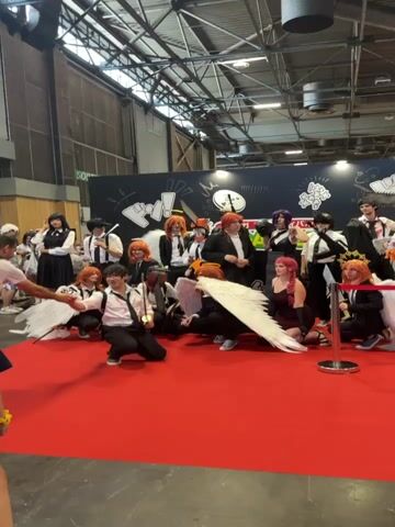 A group of cosplayers dressed as characters from Chainsaw Man are posing for a photo on a red carpet in front of a black backdrop. The backdrop has white anime-style drawings and text in Japanese. Several of the cosplayers have orange hair and wear black suits with white shirts and ties. Some of the cosplayers are holding props, including a chainsaw, a sword, and angel wings. One cosplayer in the front left is kneeling with a sword. Another cosplayer in the front center is also kneeling with a sword and large white wings. Several other cosplayers are standing and kneeling behind them. The environment appears to be an indoor convention or event space.