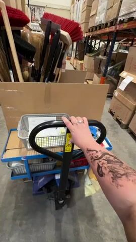 The video shows a person's arm with a large tattoo and a ring on their finger holding the handle of a pallet jack. The pallet jack is loaded with several items, including a cardboard box filled with brooms and cleaning supplies. Behind the pallet jack, there are shelves stacked with boxes in what appears to be a warehouse or storage facility. The camera is positioned from the perspective of the person holding the pallet jack, looking forward.