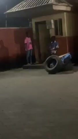 The video shows two young people standing outside a small building at night. One is wearing a pink shirt and jeans and is standing with their arms crossed. The other is wearing a blue and white patterned shirt and is holding something in their hands. A pile of tires sits in front of them. The ground is paved and there is a wall with orange paint behind the person in the pink shirt. The building has a small window and a door, and there's a shadow of someone else on the wall. The scene appears to be dimly lit.
