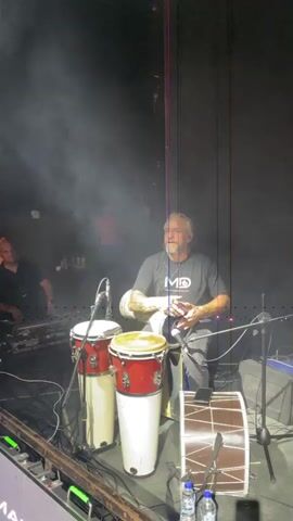 A man with graying hair and a beard, wearing a black t-shirt with a white logo and dark pants, is playing a Middle Eastern drum. He is seated and illuminated by stage lighting, with smoke or fog in the air. In front of him are two conga drums, one red and one yellow, and another larger drum with a brown and white striped pattern. To his left, another person is visible, partially obscured, operating what appears to be sound equipment with many knobs and buttons. In the background, another person in a red shirt is standing in the smoky haze. The scene appears to be on a stage, with a dark backdrop. There are microphones and cables scattered around the drums. Two plastic water bottles are on the floor in the foreground.