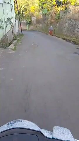 A person on a scooter rides down a paved road lined with a wall and trees. The person is wearing a helmet and a blue headscarf. A second person on a scooter follows behind. The second person is wearing a backpack. The video is shaky, suggesting it was filmed from a moving vehicle. The sky is bright, indicating it is daytime. The road appears to be in a rural or suburban area.