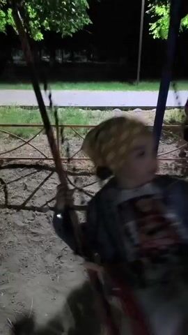 A young child with blonde hair wearing a t-shirt and shorts is on a swing set at night. The child is swinging back and forth. The swing set is on a dirt ground and there is a metal fence behind the swing. There are trees in the background and the area is lit by artificial light.
