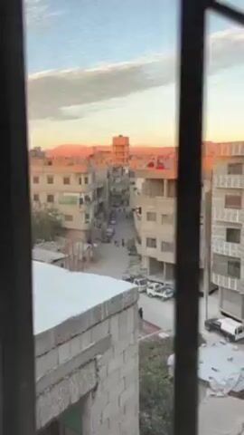 The video depicts an urban street scene, viewed from an elevated position, possibly a window. The street is lined with multi-story buildings constructed from light-colored materials, likely concrete or brick. The buildings show signs of wear and some appear to be under construction or incomplete. In the background, there are mountains faintly visible against a sky tinted with the warm hues of sunrise or sunset. 

The street below is narrow and cluttered with parked cars. A motorcycle is seen driving down the center of the street, along with several pedestrians, including two children in blue jackets, a person wearing a hijab and carrying a bag, and a person wearing what looks like a winter jacket. The street is crisscrossed with overhead wires. A truck with green foliage attached to it is parked to the right.