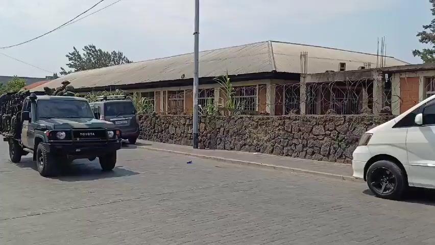 A military vehicle, a Toyota Land Cruiser pickup truck painted in camouflage, is driving down a paved street. The truck bed is filled with soldiers in full military gear, including helmets, flak jackets, and rifles. They are seated and facing forward. To the side of the military vehicle is a white minivan. A building with a stone wall and barbed wire sits in the background.
