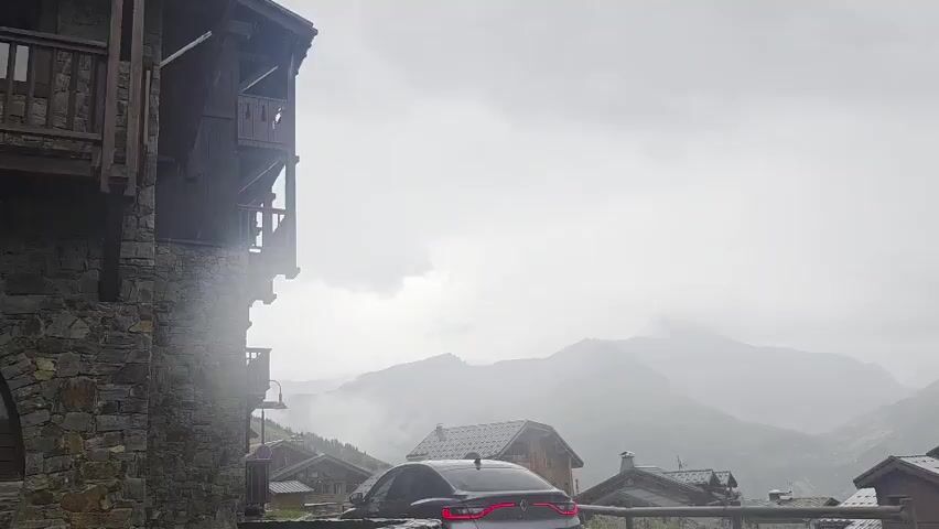 The video shows a mountain village on a cloudy day. In the foreground, a dark car is parked on a wet road. Traditional wooden houses with dark roofs are visible on either side of the road. In the background, misty mountains rise up, partially obscured by clouds. The overall atmosphere is somewhat dreary and atmospheric, suggesting a cool, damp climate.