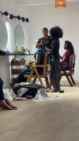 The video shows a beauty salon with three women getting ready for something. One woman is sitting in a chair while two others stand behind her, working on her hair. Another woman is sitting in a chair in the background. In the foreground, another woman is sitting on a couch while two others kneel in front of her, near a light on a tripod and equipment. All of the women are Black, and the women working on others are likely hair or makeup stylists. The room is decorated with round mirrors, wicker baskets, and decorative lights. There are plastic bags and equipment on the floor, suggesting they are in the middle of preparations.