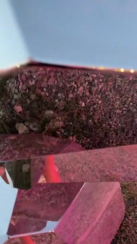 The video shows a faceted mirrored sculpture from a low-angle perspective. The sculpture is outdoors, with gravel and grass visible around its base. The sky is visible in the upper portion of the frame, with some clouds. Reflections in the mirrored surfaces show parts of the environment, including trees, lights, and people. One reflection clearly shows a person holding a phone and taking a selfie. Another reflection shows a person's legs and feet. The lighting appears to be natural daylight, with some pink and blue colored lights from within or around the sculpture. The camera is moving, panning and tilting to capture different angles of the sculpture and its reflections.