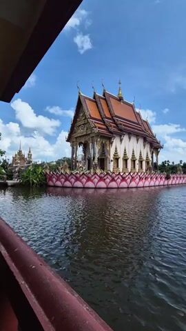 The video presents a scenic view of a traditional Thai temple, which is partially submerged in water, possibly a lake. The temple has a distinctive orange and white color scheme and a multi-tiered roof structure common in Thai architecture. Notably, the base of the temple appears to be decorated with pink lotus-shaped structures, adding to its visual appeal. The water surrounding the temple is calm with slight ripples. The sky overhead is blue with scattered white clouds, indicating a clear and sunny day. The shot appears to be taken from a boat, moving slightly, as suggested by the visible edge of the boat and the changing perspective of the temple against the background.