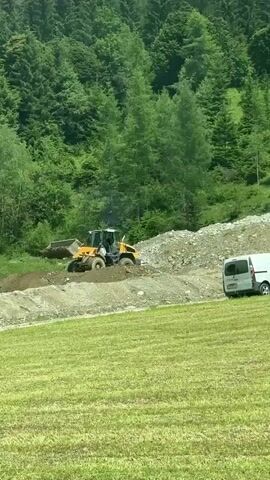 This media file captures a front-end loader, a piece of heavy construction equipment, operating on a hillside. The machine is yellow and black and appears to be actively moving dirt or gravel. The surrounding environment is a steep, tree-covered mountainside, characteristic of a mountainous region. The trees are lush and green, suggesting a temperate season, likely spring or summer. The weather appears to be overcast or hazy, as there are no harsh shadows and the light is diffused.

The loader is positioned on a graded area of the hill, with piles of excavated material visible. The foreground shows a grassy slope, possibly a field or meadow, which is a lighter shade of green. A dirt track or road runs horizontally across the midground, partially obscured by the loader and the disturbed earth. The overall scene suggests some form of construction or land management activity is underway in a rural, mountainous setting.

The image is a still frame taken from a video, indicated by the timestamp "00:00" in the top left corner. The media appears to be captured during daytime, given the ambient light. The location context provided is Flachauwinkl, Austria, which aligns with the mountainous terrain and dense forestation depicted. There is no visible text within the frame itself. The activity is focused on the machinery and its task, with no people visible. The overall mood is one of industrious activity within a natural landscape.