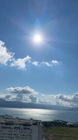 The video shows a bright sunny day with a clear blue sky. The sun is high in the sky and casts strong rays. There are some white clouds scattered across the sky. Below the sky, there is a body of water, likely a sea or ocean, with waves reflecting the sunlight. In the foreground, there is a large apartment building with many windows, suggesting an urban or suburban environment. Further in the distance, there are rolling hills or mountains. The overall scene depicts a serene landscape with a vibrant sky and a glimpse of coastal life.