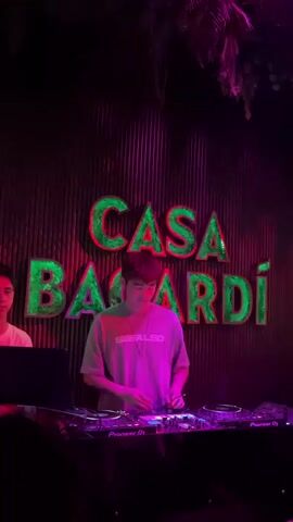A DJ, wearing a pink t-shirt and dark pants, stands behind a DJ booth and mixes music in front of a dark corrugated wall with a large "CASA BACARDI" sign illuminated in green glitter. The DJ booth is equipped with two Pioneer CDJ-2000 Nexus turntables and a Pioneer DJM-900 Nexus mixer. The scene is lit with pink and purple stage lights, creating a vibrant atmosphere in what appears to be a nightclub or event space. The DJ is focused on the equipment, with their hands on the turntables. A second person, wearing a white t-shirt, is partially visible to the left of the DJ, also looking towards the DJ booth. The overall impression is one of a lively nighttime event.