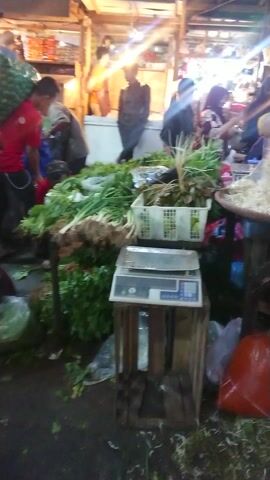 The video captures a bustling market scene, likely at night or in the very early morning, evident from the prominent artificial lighting that casts bright gleams and shadows. Numerous people are visible, moving through the market either as shoppers or vendors, contributing to a lively atmosphere.

Market stalls are densely packed with a variety of fresh produce. In the initial frames, a stall features baskets holding green leafy vegetables, possibly spring onions, alongside a larger basket containing what appears to be raw noodles or thin pasta. Nearby, yellow fruits, resembling lemons or small gourds, are piled high, and round, brownish fruits (possibly salak) are also displayed. Further into the video, large woven baskets are filled with long, green, elongated vegetables, which could be long beans or petai (stink beans). Another prominent display showcases several large baskets overflowing with dark, oval-shaped items, strongly suggesting shellfish such as mussels or clams.

A vendor, wearing a light-colored cap and a grey shirt, is seen behind one of the stalls filled with the dark oval items. A digital scale is positioned on this stall, indicating that goods are being weighed for customers. Throughout the footage, individuals are seen navigating the crowded pathways, some carrying bags, reinforcing the image of an active and busy marketplace. The ground appears somewhat wet and uneven, characteristic of a fresh food market. The scene depicts a vibrant commercial exchange in what appears to be a covered market area in Coubron, France. No discernible text is visible on any signs or products.