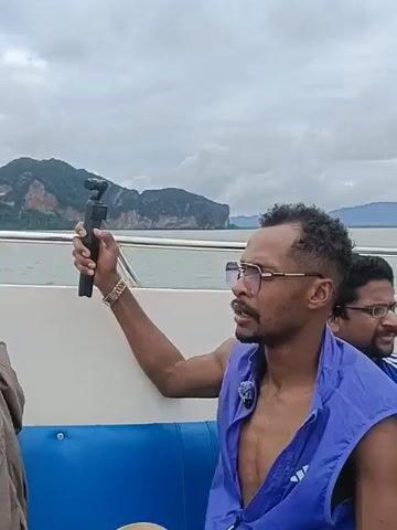 The video shows a group of people on a boat. In the first frame, a man with dark curly hair and glasses is holding a camera on a selfie stick, looking to his right. He is wearing a blue vest over his bare chest and a watch on his left wrist. In the background, there are islands and a cloudy sky. The boat appears to be moving through the water, as indicated by the movement of the camera and the scenery. The second frame shows a woman with blonde hair facing away from the camera, looking at the scenery. The third and fourth frames show the view from the front of the boat, with more islands and the sea visible. The overall environment appears to be a tropical or subtropical location with a body of water and mountainous islands.