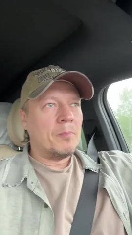 A man is sitting in a car. He is wearing a baseball cap, a light green button-down shirt, and a beige t-shirt. He has short, blonde hair and a light beard. He is wearing a seatbelt. The car interior is gray. The man is looking at the camera and singing. The background is out of focus, but appears to be a road with trees.
