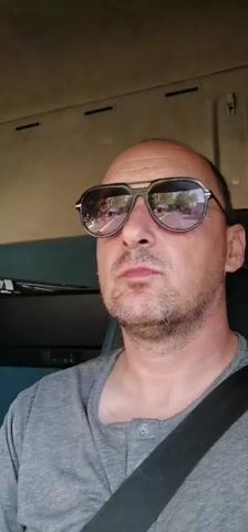 A man wearing sunglasses and a gray shirt is driving. He appears to be in a truck. A seatbelt is visible across his chest. Music plays.