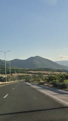 This is a point-of-view shot from a vehicle traveling on a highway under a clear blue sky. The road curves to the left, and on the right, there is a metal guardrail separating the highway from a vegetated area with scattered trees and fields. In the distance, a large, tree-covered mountain range dominates the horizon. Streetlights are visible along the side of the highway. The time stamp indicates the beginning of the video.