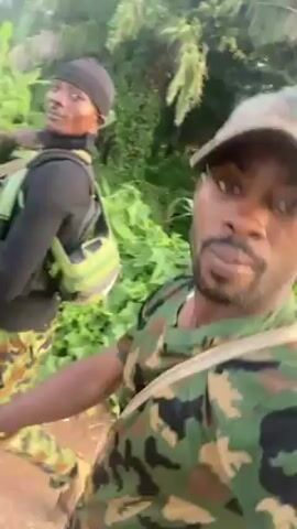 Two men wearing camouflage clothing are walking down a dirt road in a wooded area. The man in the foreground is wearing a camouflage hat and carrying a rifle. The man in the background is wearing a black beanie and a black vest over his camouflage clothing. He is also carrying a rifle. The man in the foreground is talking to the camera, while the man in the background is making a hand gesture.
