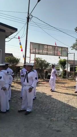 A group of men in white naval uniforms are gathered outdoors. Some are standing in a line, while others are conversing. In the background, there is a building, a sign with Indonesian writing, trees, and a clear blue sky. A microphone stand is visible in the foreground. The men appear to be participating in some kind of official event or ceremony.