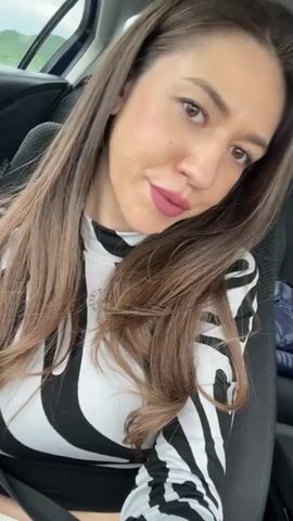 The video shows a woman sitting in the back seat of a car. She has long brown hair and is wearing a black and white zebra-print top. She is smiling at the camera. There is a jacket lying on the seat next to her. The background is blurred, but it appears to be daytime. The camera is slightly angled down towards her. There is music playing in the background.