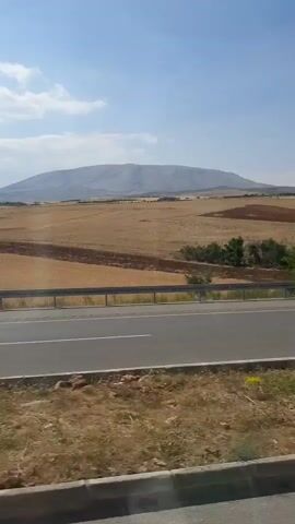 The video shows a view from a moving vehicle. A highway with two lanes is visible in the foreground. Beyond the highway, there are fields of dry grass and some trees. In the background, there is a large mountain range under a clear blue sky with a few scattered clouds. The camera is capturing the scenery as the vehicle travels along the road. There are no people or other vehicles clearly visible in the frame.