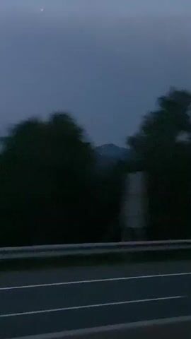 The video shows a blurry, dark landscape from the perspective of a moving vehicle. In the foreground, there is a dark asphalt road with white lines. A metal guardrail separates the road from the terrain beyond. The middle ground features dark green trees and foliage, and in the background, there are dark, rolling hills or mountains. The sky is a deep, dusky blue. There are no people or discernible actions visible. The overall impression is of a nighttime or twilight journey through a rural or mountainous area.