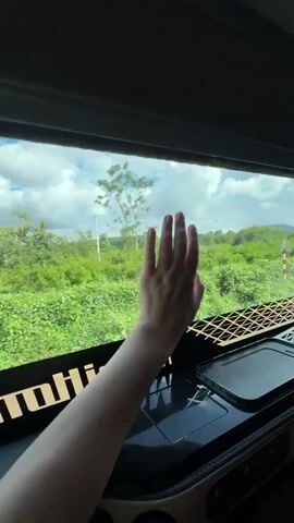 A first-person perspective shot from inside a vehicle, looking out a window at a lush green landscape with a partly cloudy sky. A person's left arm and hand are visible in the foreground, with their hand raised and resting against the window. The arm and hand are fair-skinned. Below the window is a dark dashboard with a textured metal grate and a black smartphone resting on it. The vehicle appears to be in motion, as evidenced by the slight blur of the scenery outside. The text "MohidM" is partially visible on the dashboard.