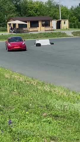 A red Tesla Model 3 is performing maneuvers on an asphalt track. The car is observed from multiple angles as it navigates around orange traffic cones and white barriers. The environment appears to be an outdoor facility, possibly a race track or driving school, with grass and trees bordering the track. The sky is partly cloudy. There are no visible people outside of the driver inside the Tesla. The car is in motion throughout the video, demonstrating acceleration, cornering, and possibly braking.