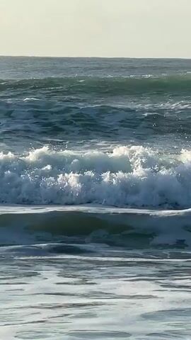 A close-up, eye-level shot captures the dynamic motion of ocean waves breaking on a beach. The water is a mixture of deep blue and vibrant turquoise, with white foam cresting and churning as the waves roll towards the viewer. The sunlight illuminates the scene, creating a bright, hazy sky in the background. The waves are layered, with smaller waves in the foreground and larger, more powerful waves in the mid-ground and background, all moving in the same direction. There are no people or objects visible in the frame, emphasizing the raw power and beauty of nature. The overall impression is one of constant movement and energy, a serene yet forceful display of the ocean's might.