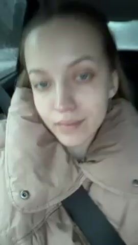 A woman with light skin and light hair pulled back is sitting in the passenger seat of a car wearing a beige puffer jacket and a seatbelt. She is looking at the camera with a slight smile. The background shows a window with raindrops, suggesting it's raining outside. The car interior is dark.