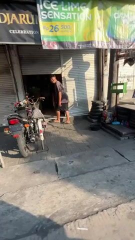 A person in a grey shirt and black pants is bent over near the entrance of a closed shop with a metal roll-up door. A bicycle with a basket is parked to the left. To the right, stacked tires and various objects are visible on the ground next to a pole. The scene appears to be an outdoor urban environment with a concrete surface and shadows cast by the sun.